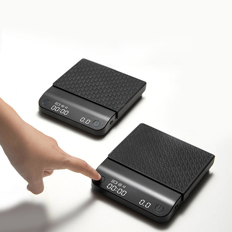Black coffee electronic scale with textured top, buttons, and hand reaching toward it